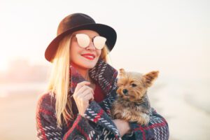 Portrait of beautiful smiling hipster trendy blonde woman walking on the beach with a little dog wearing sunglasses and black hat