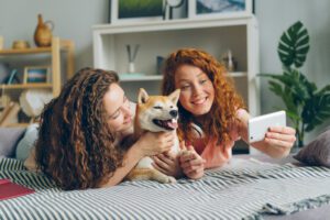 Siblings beautiful girls are taking selfie with cute puppy lying on couch at home using smartphone camera. Modern technology, people and animals concept.